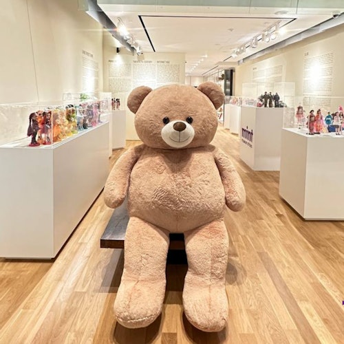Large tan teddy bear seated on a bench in a museum-style display area filled with glass cases showcasing various dolls.