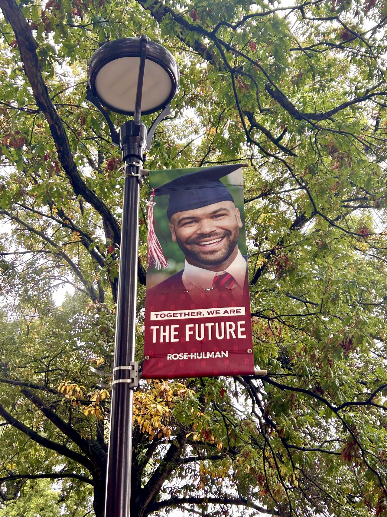 A streetlight with a banner showing a smiling graduate wearing a cap and gown. The banner reads, "Together, we are the future – Rose-Hulman." The background features green foliage from the trees above.