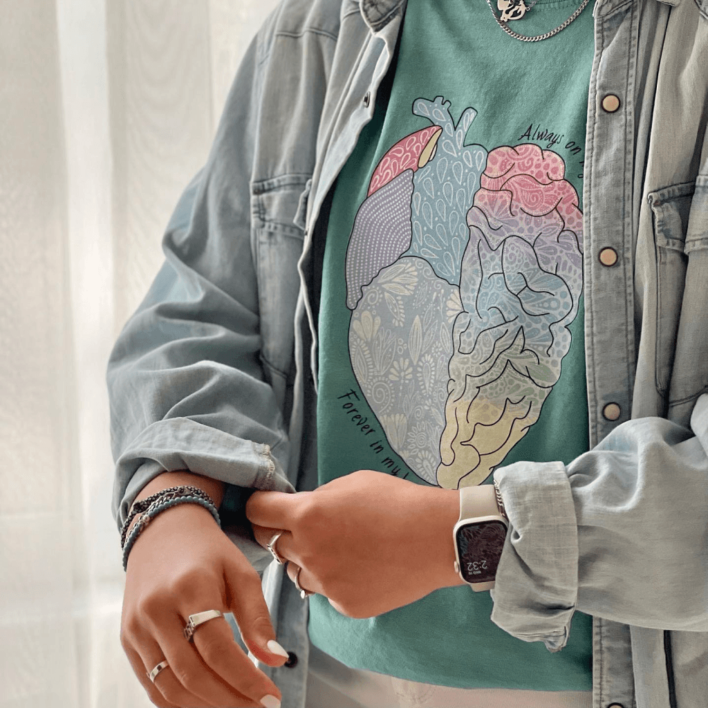 A person wears a stylish, eco-friendly shirt featuring a colorful anatomical heart design, complemented by a light denim jacket and layered silver necklaces.