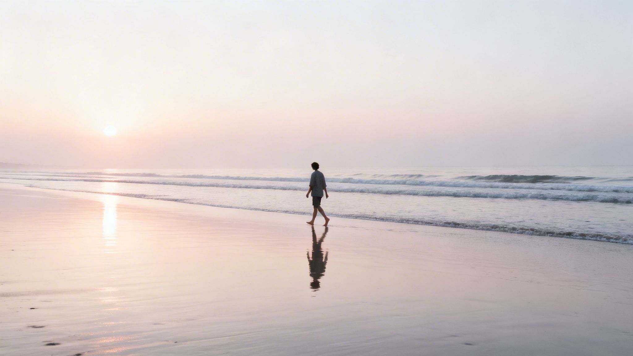 A person walking barefoot on a tranquil beach at sunrise, reflected on the wet sand.
