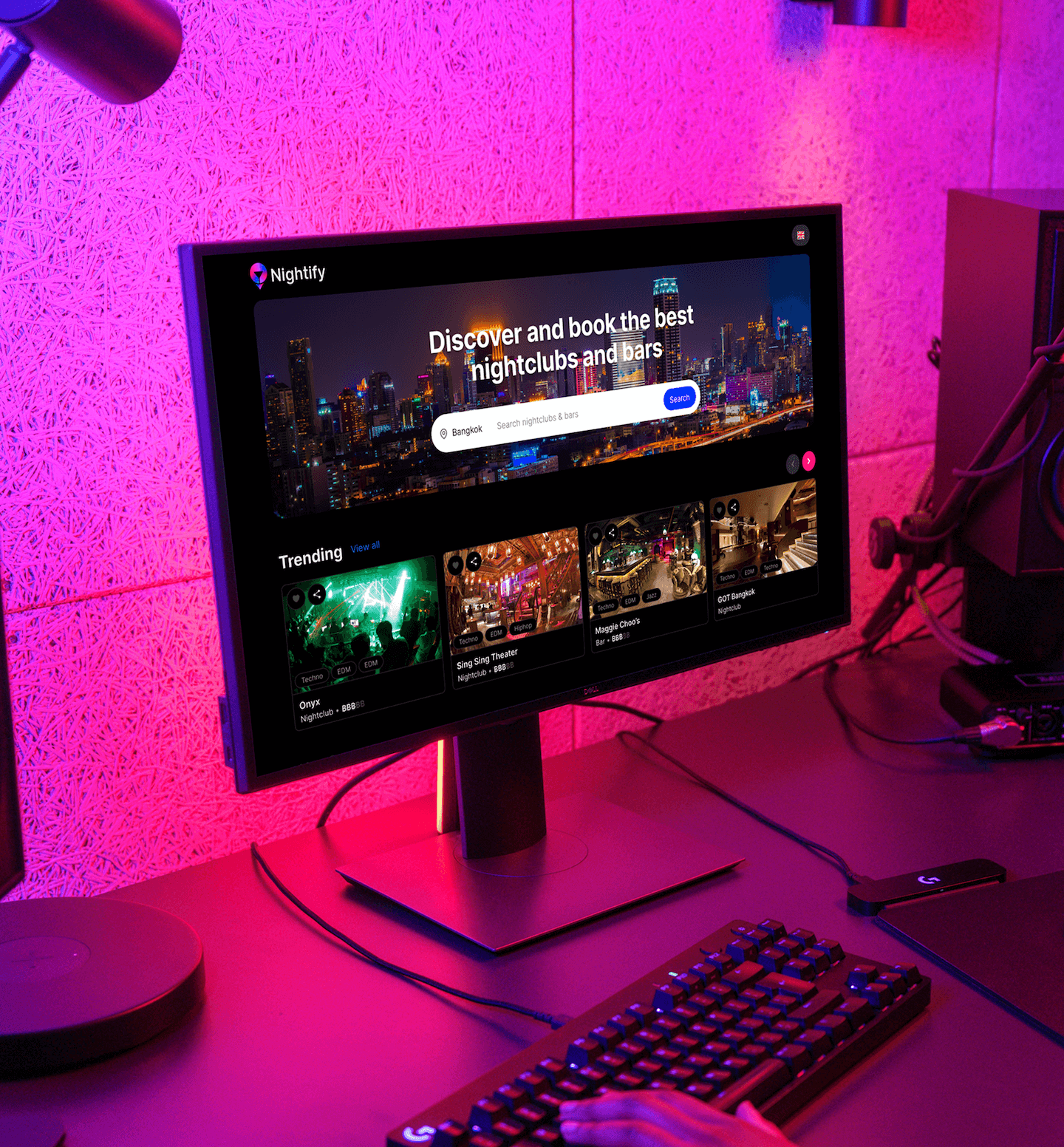 Desktop UI design mockup of the Nightify web platform displayed on a monitor in a professional workspace. The interface features a prominent "Discover and book the best nightclubs and bars" search bar over a high-contrast city skyline background.