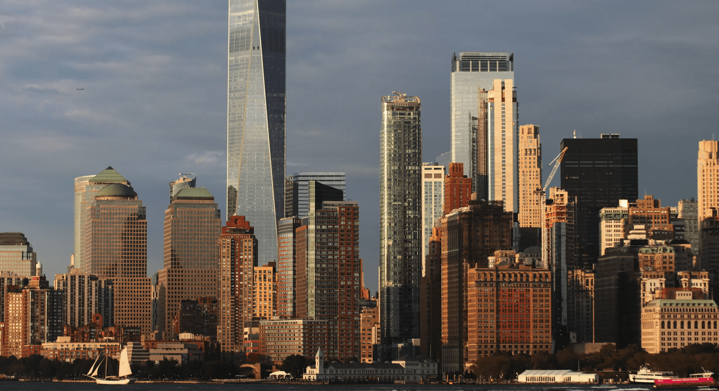 View of the Lower Manhattan skyline with One World Trade Center and surrounding skyscrapers illuminated by warm evening sunlight.