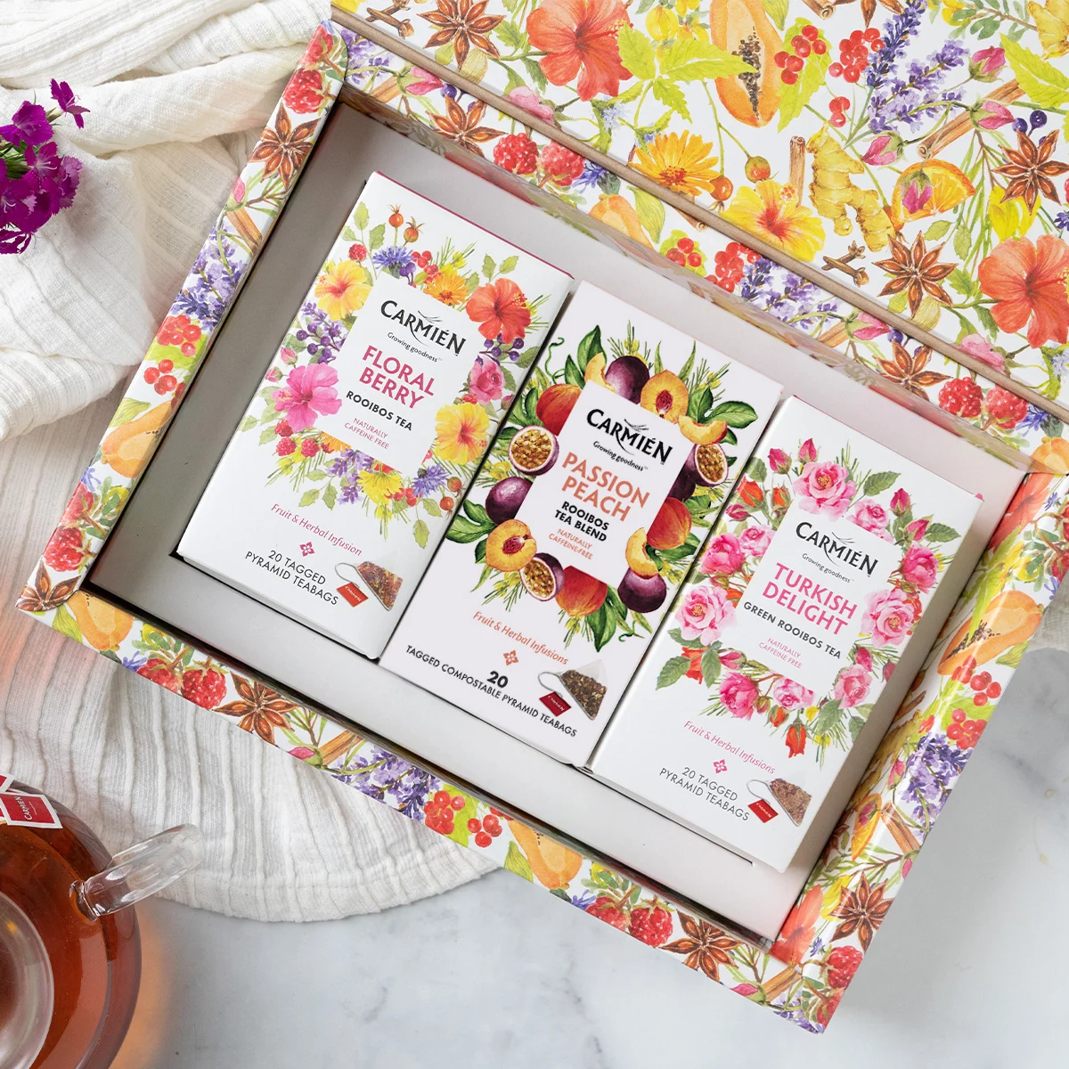 An open gift box containing an assortment of Carmién tea blends in small boxes, with a full glass tea kettle partially in view from the bottom-left corner of the composition.