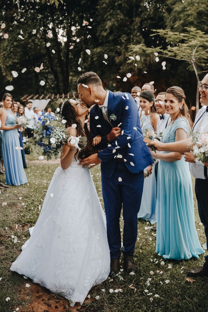 Newlyweds sharing a kiss as white rose petals rain down surrounded by cheering guests in blue dresses at an outdoor garden wedding