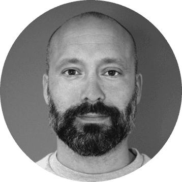 Black and white circular portrait of a man with a beard and short hair looking directly at the camera.