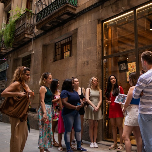 Skupina od osam osoba stoji na uskoj ulici ispred trgovine s otvorenim staklenim vratima i razgovara.