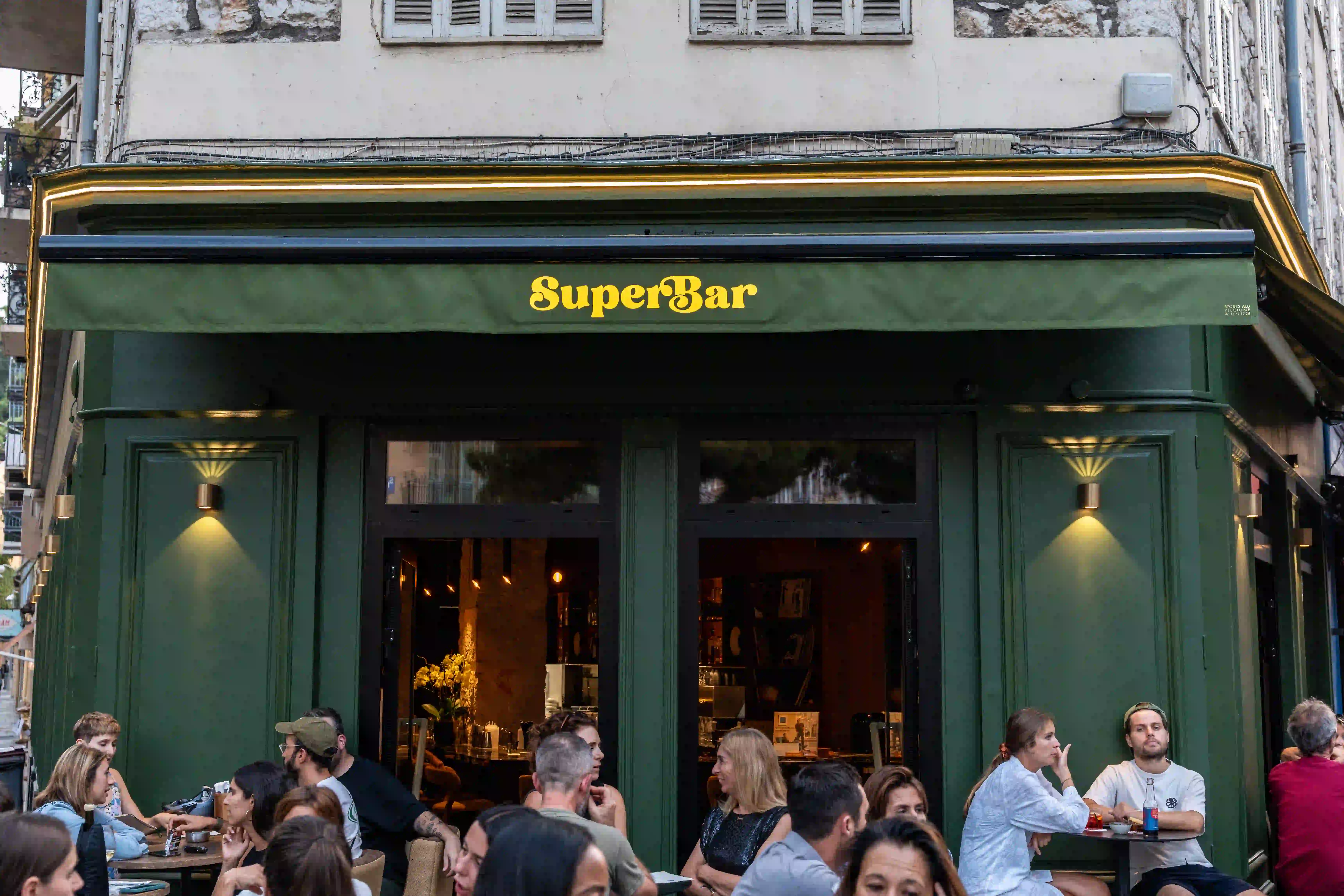 A bustling outdoor café scene with people casually seated at tables in front of "SuperBar," featuring a green canopy and warm ambient lighting on a European city street.