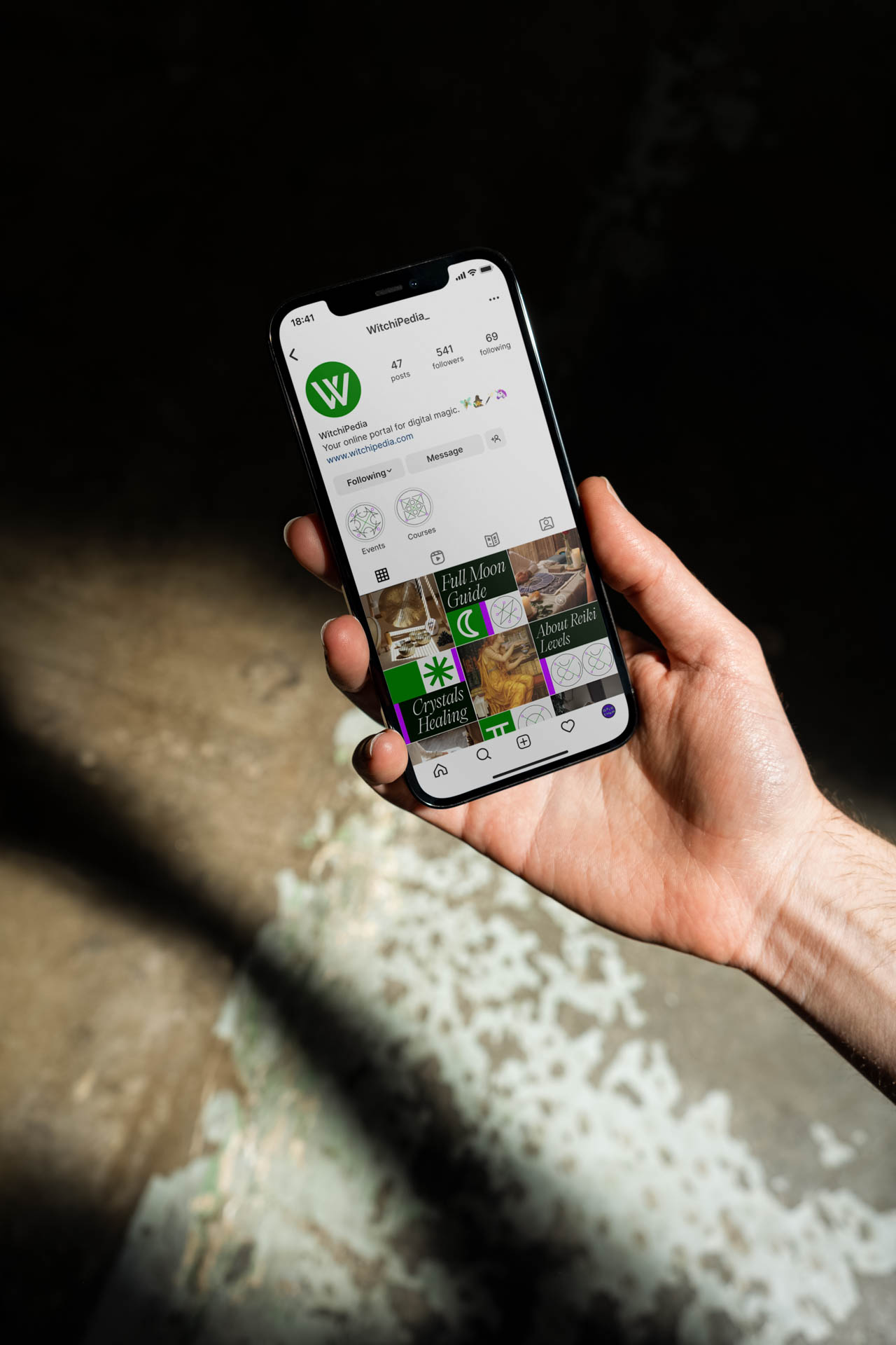 A hand holds a smartphone displaying the Witchipedia app interface. The screen shows search results for "Witchipedia" with a green "W" logo. The phone is held against a dark background with a light-colored surface visible in the lower portion of the image. The phone’s screen shows various search results, including "Gothic Cuisine," "Crystals," "Hedgehogs," and "Culinary Herbs." The time displayed on the phone is 15:41.