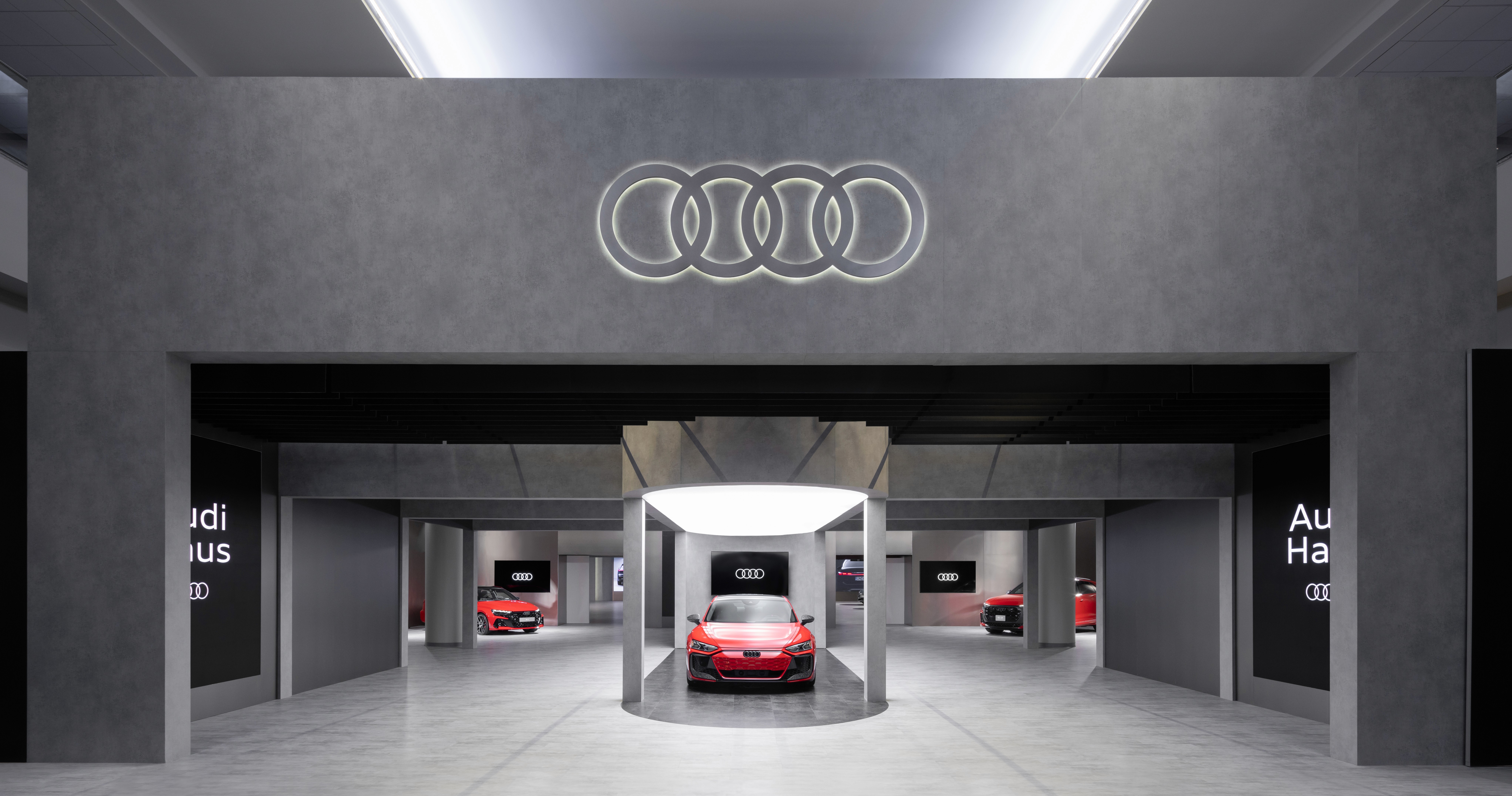 A modern showroom features a sleek red car under soft lighting in the center. The Audi logo glows above. Minimalist design with concrete textures.