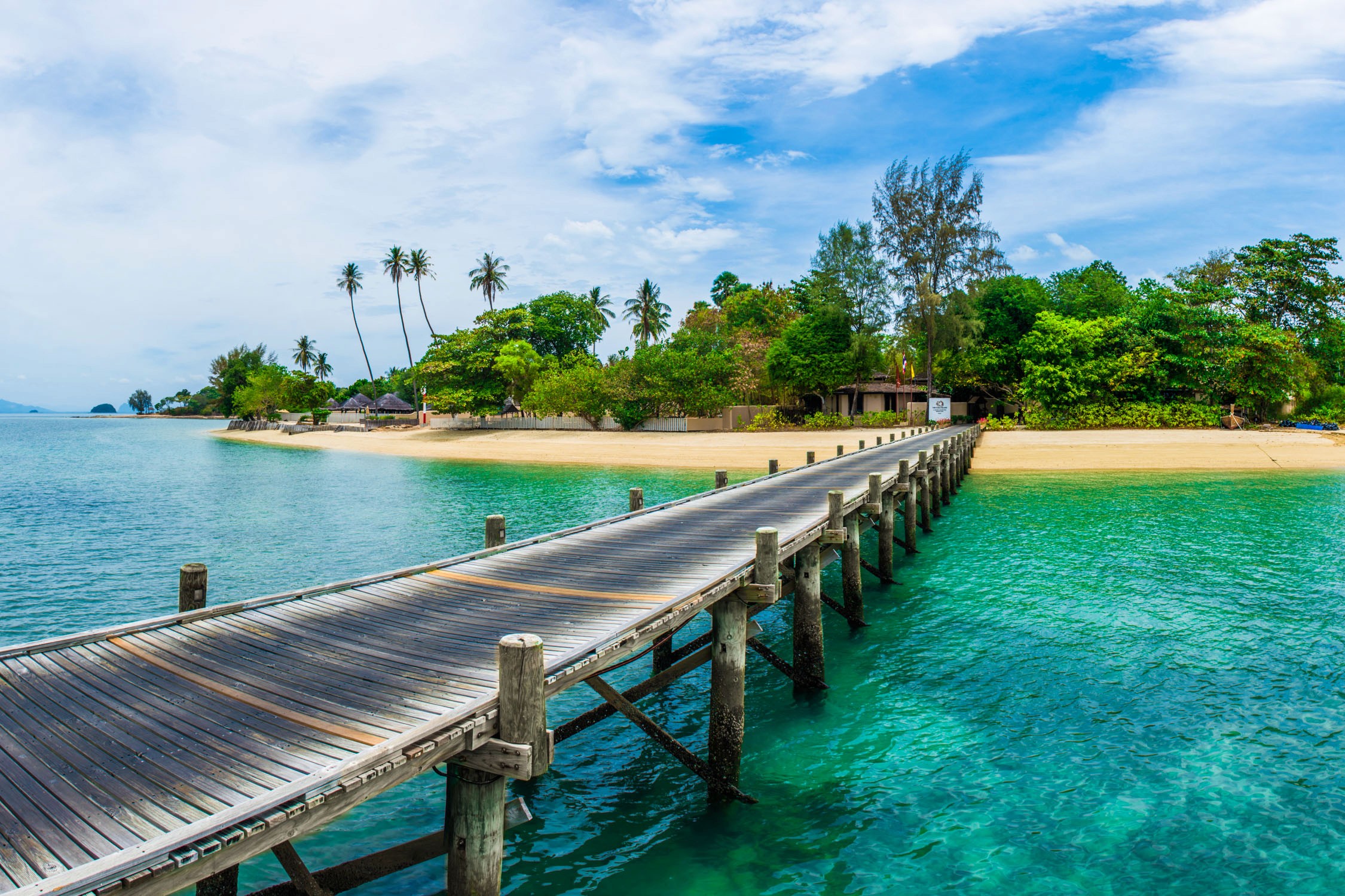 A wooden pier extends over turquoise water toward a tropical beach lined with palm trees and lush green foliage.