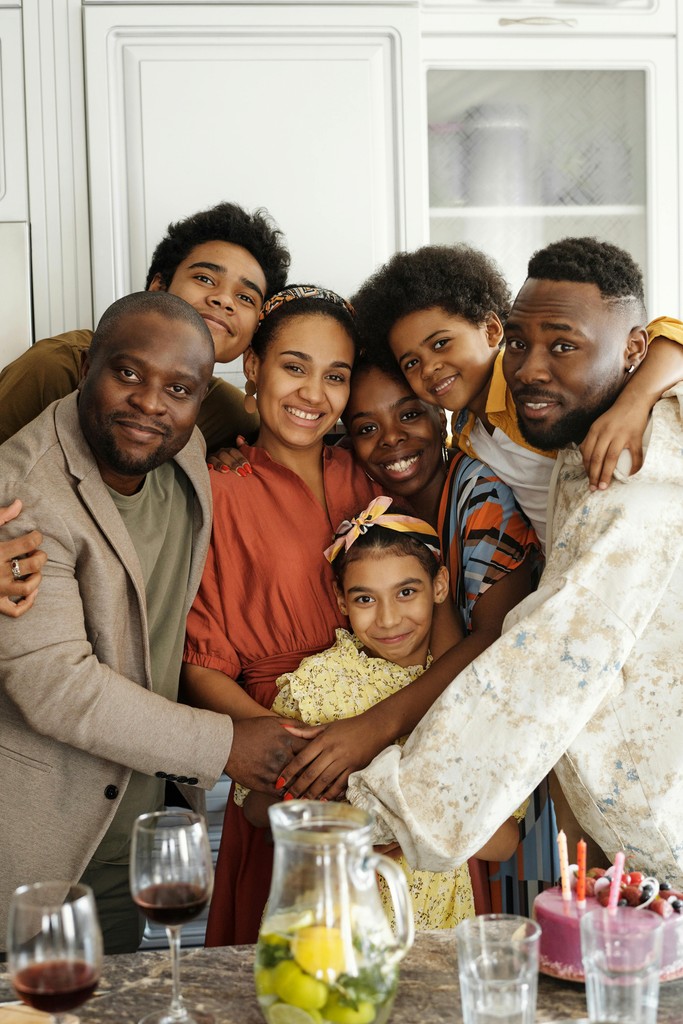 The image captures a joyful and close-knit group of six people, including adults and children, all smiling and embracing each other. The diverse family or group of friends radiates warmth and happiness, highlighting the strong bonds and affection they share. The setting appears to be indoors, with everyone dressed in colorful and casual attire, enhancing the lively and heartwarming atmosphere.