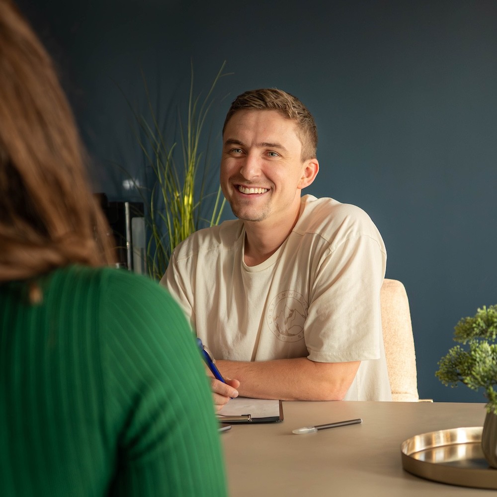 Zwei Personen sitzen sich an einem Tisch gegenüber, eine Person hält einen Stift und ein Klemmbrett