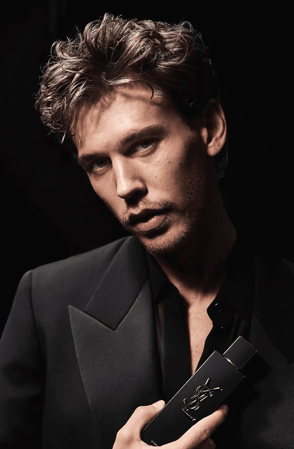Austin Butler in a black suit, posing with an intense expression while holding YSL Myslf fragrance.