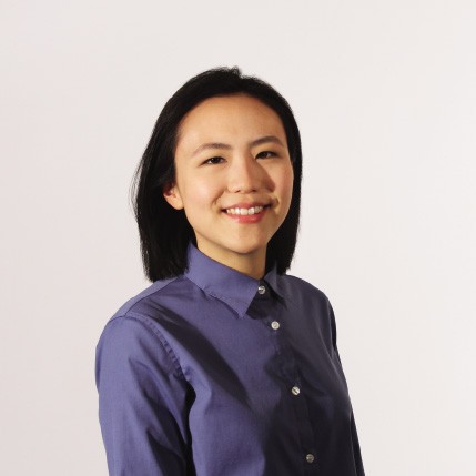 A person with short dark hair is captured in a head-and-shoulders portrait against a plain off-white background. They are wearing a dark blue, button-down shirt with long sleeves and a standard collar. The individual is facing forward and smiling slightly, with their gaze directed straight at the camera. The lighting is even and soft, casting minimal shadows. The image is well-lit and focuses primarily on the person's face and upper body. No other objects or text are visible in the frame.