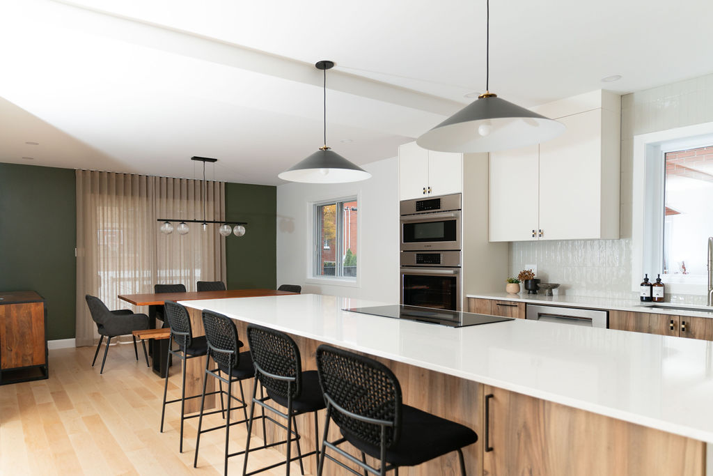 Cuisine fini blanche et bois. Four encastré au mur. Réfrigérateur sur le mur du fond. Une fenêtre se trouve au-dessus de l'évier. Dosseret de céramique installé à la vertical blanc lustré. Armoires blanches mates. Gros îlot entièrement en bois. Rangement intégrés avec quatre places assises. Tabouret noirs avec dossier. Comptoir en quartz blanc. Plaque de cuisson intégrée dans l'îlot. Suspension noir et blanc au-dessus de l'îlot. Plancher en bois clair. On observe un mur vert qui encadre la porte-patio et met en valeur l'habillage de la fenêtre de couleur beige. La table à manger est en bois foncé avec des chaises noires.