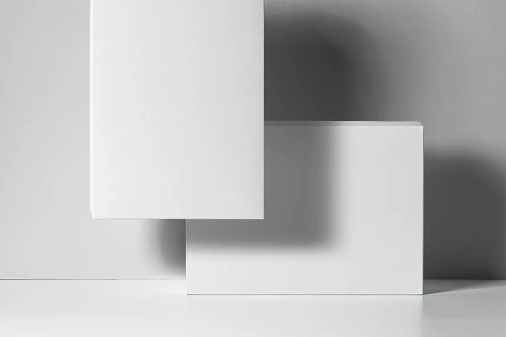 Rectangular product box mockup featuring two designs: one in black with colorful abstract shapes and bold text, and another in peach with similar graphics. Displayed against a black and white background with soft lighting.