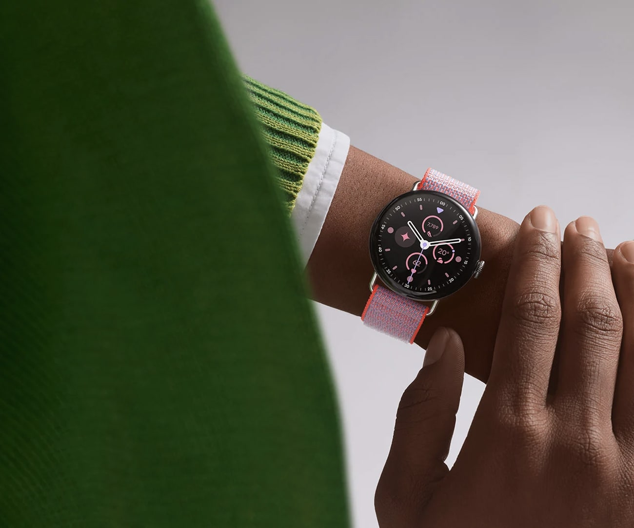 Fotografia de um pulso usando um relógio com fundo preto e pulseira rosa. A pessoa veste um casaco verde claro.