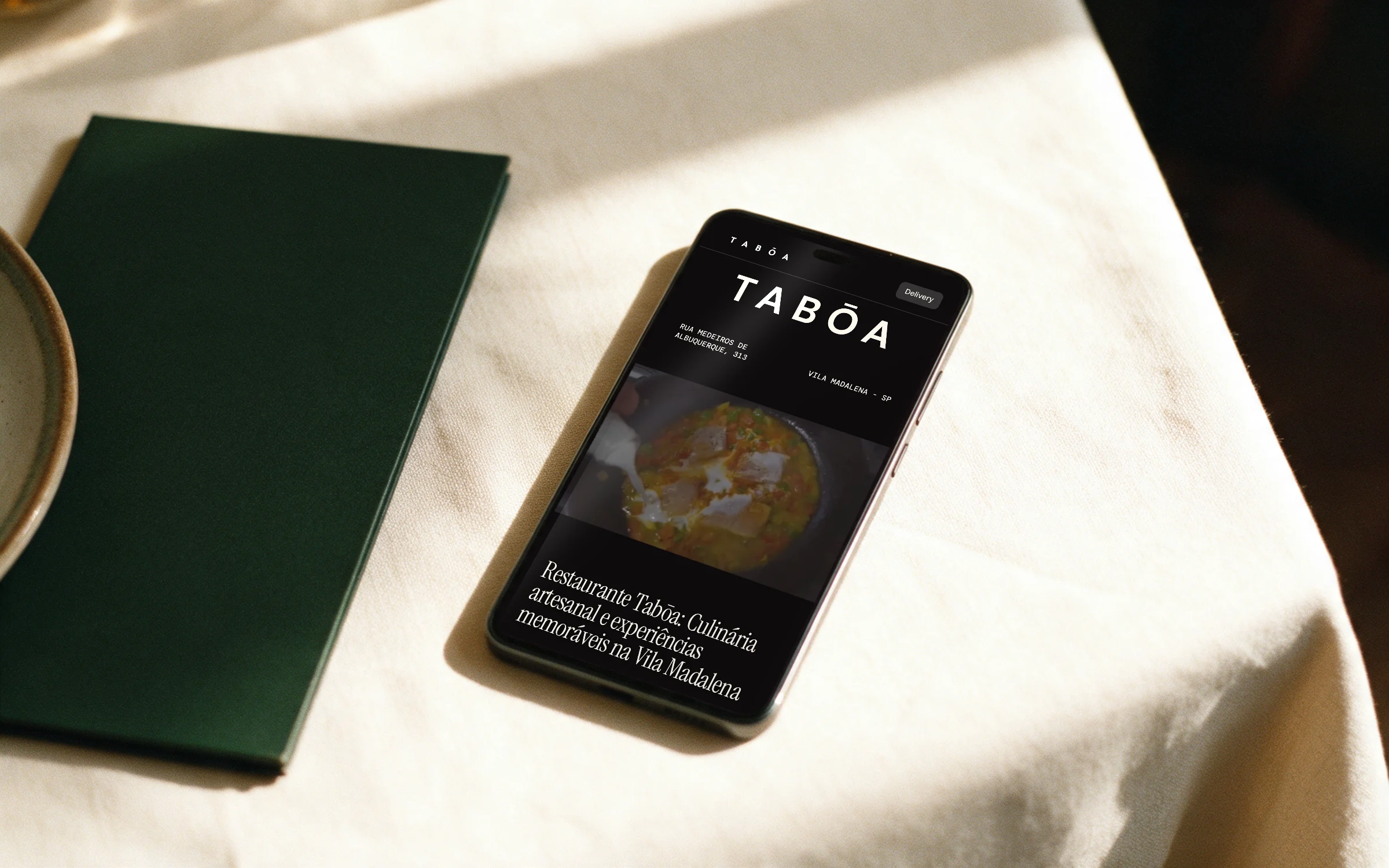 Página inicial do site do Tabōa num celular sobre uma toalha de linho