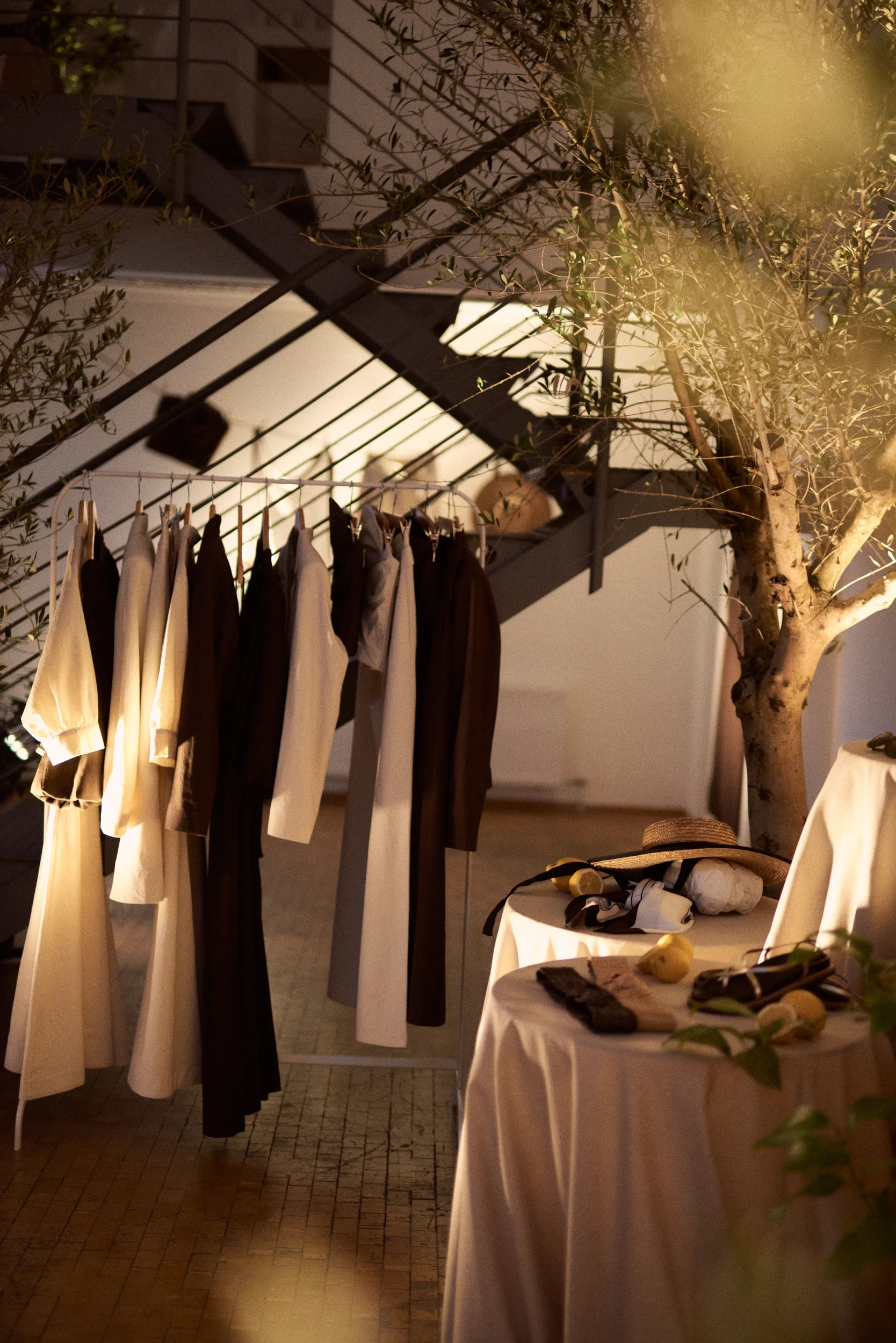 Clothing rack with cream and brown looks next to an olive tree and styled side tables.