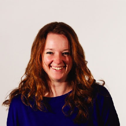 A woman with long, curly, reddish-brown hair smiles at the camera. She wears a dark blue top against a plain off-white background.