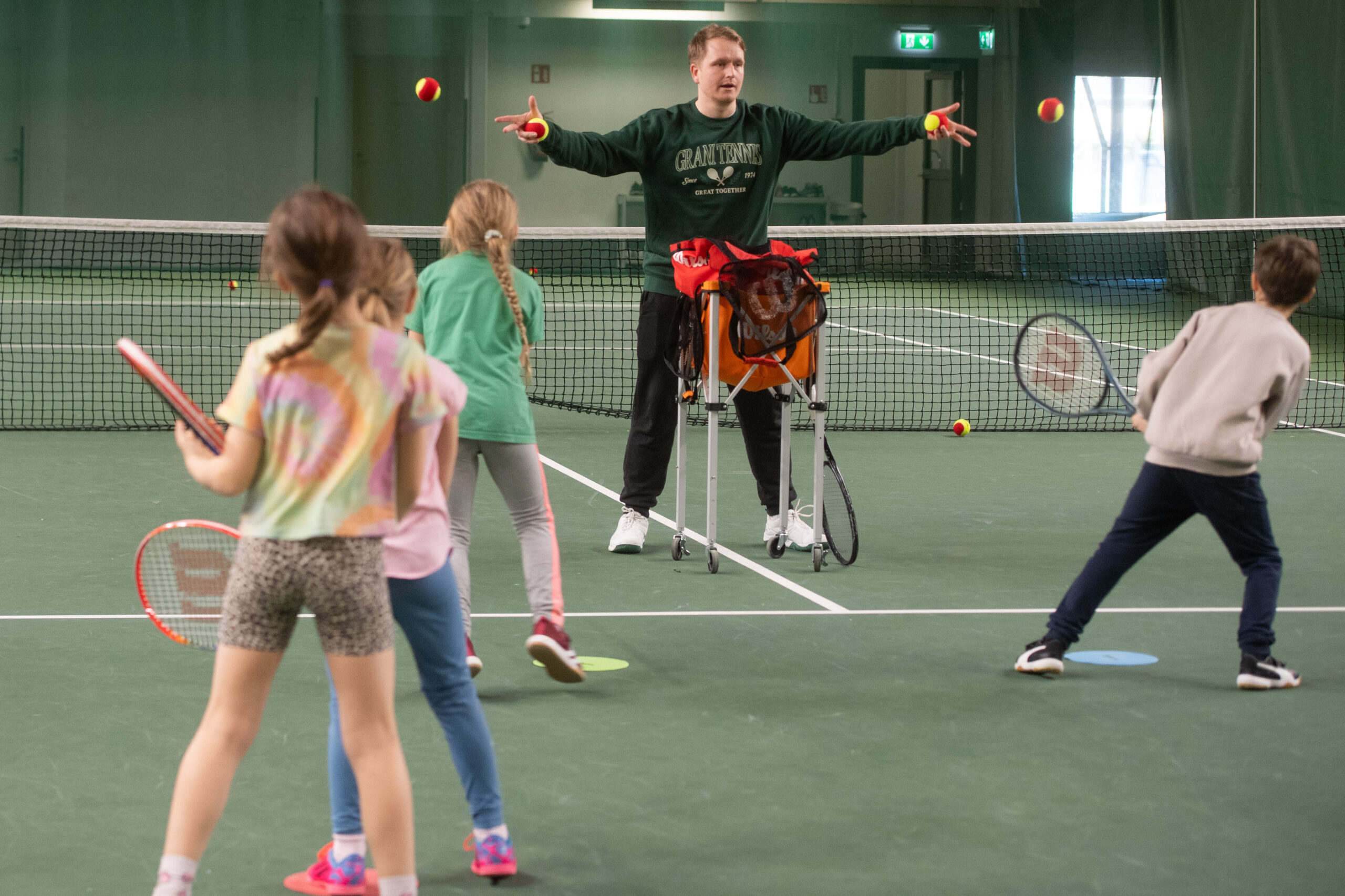 Lapset harjoittelevat tennistä valmentajan heittäessä palloja