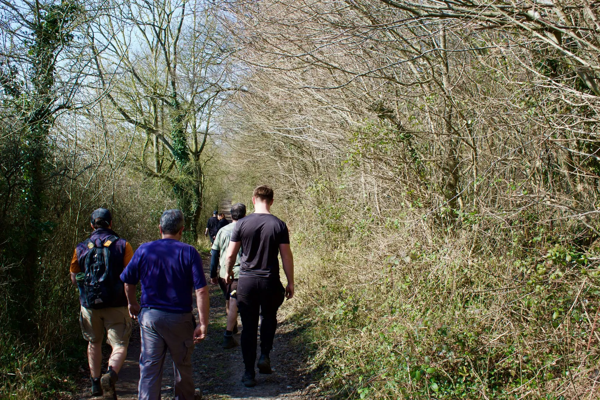 A group of people walking along a wooded path surrounded by greenery on a sunny day.