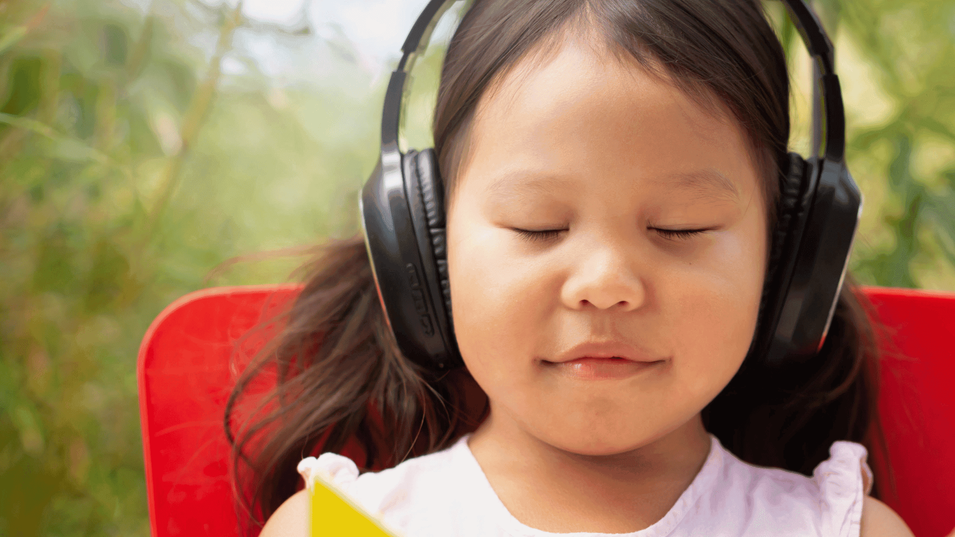 A young girl sitting on a red chair with her eyes closed, wearing headphones and looking peaceful while listening to HushAway®’s Sound Sanctuary.