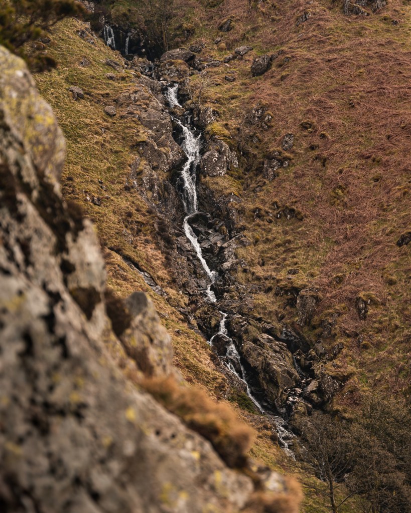 A tall waterfall cascades down the side of a fell. The photo is taken from behind a rock that's blurred in the foreground.