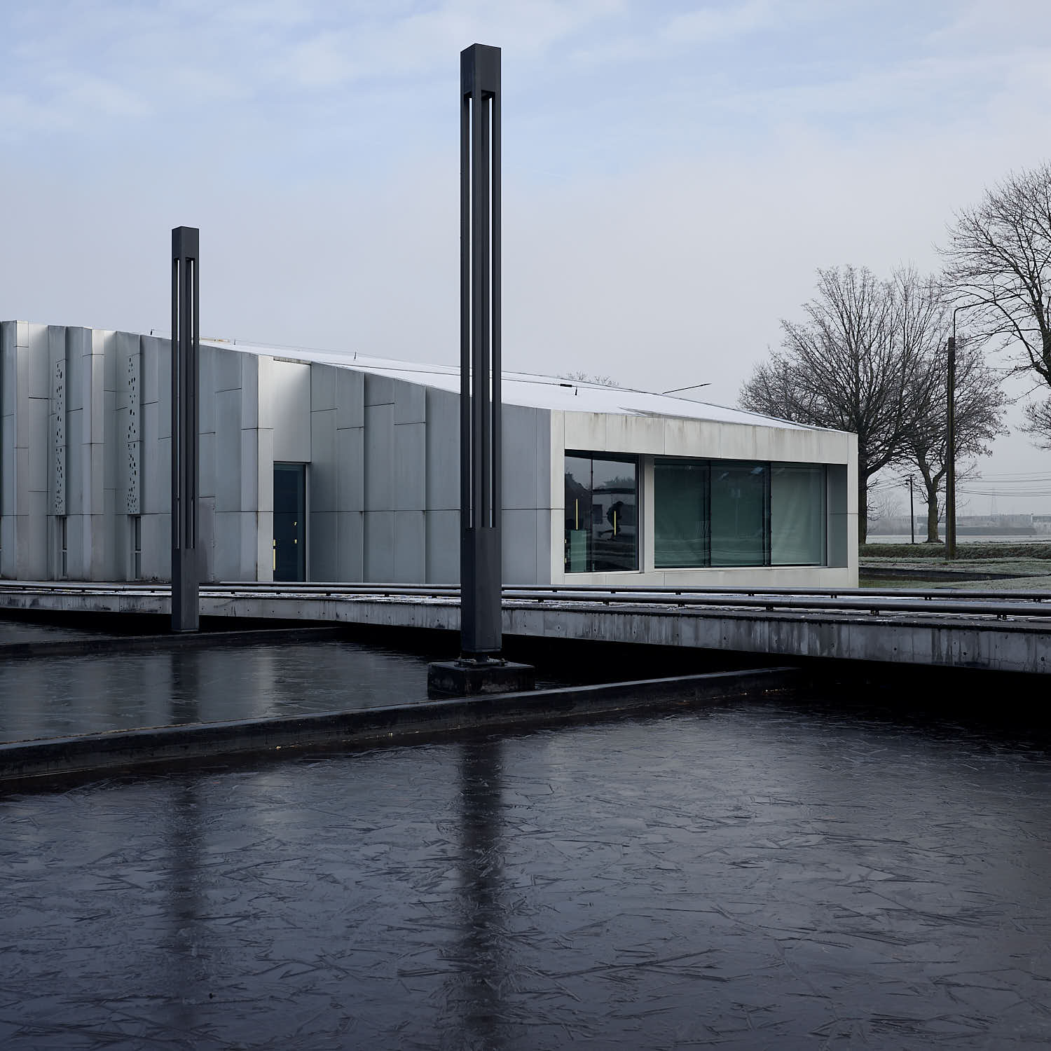 Photographie d'architecture extérieure du Centre Culturel de Lesquin : façade moderne blanche et rythmée se reflétant dans un bassin d'eau gelé sombre avec des luminaires verticaux graphiques.