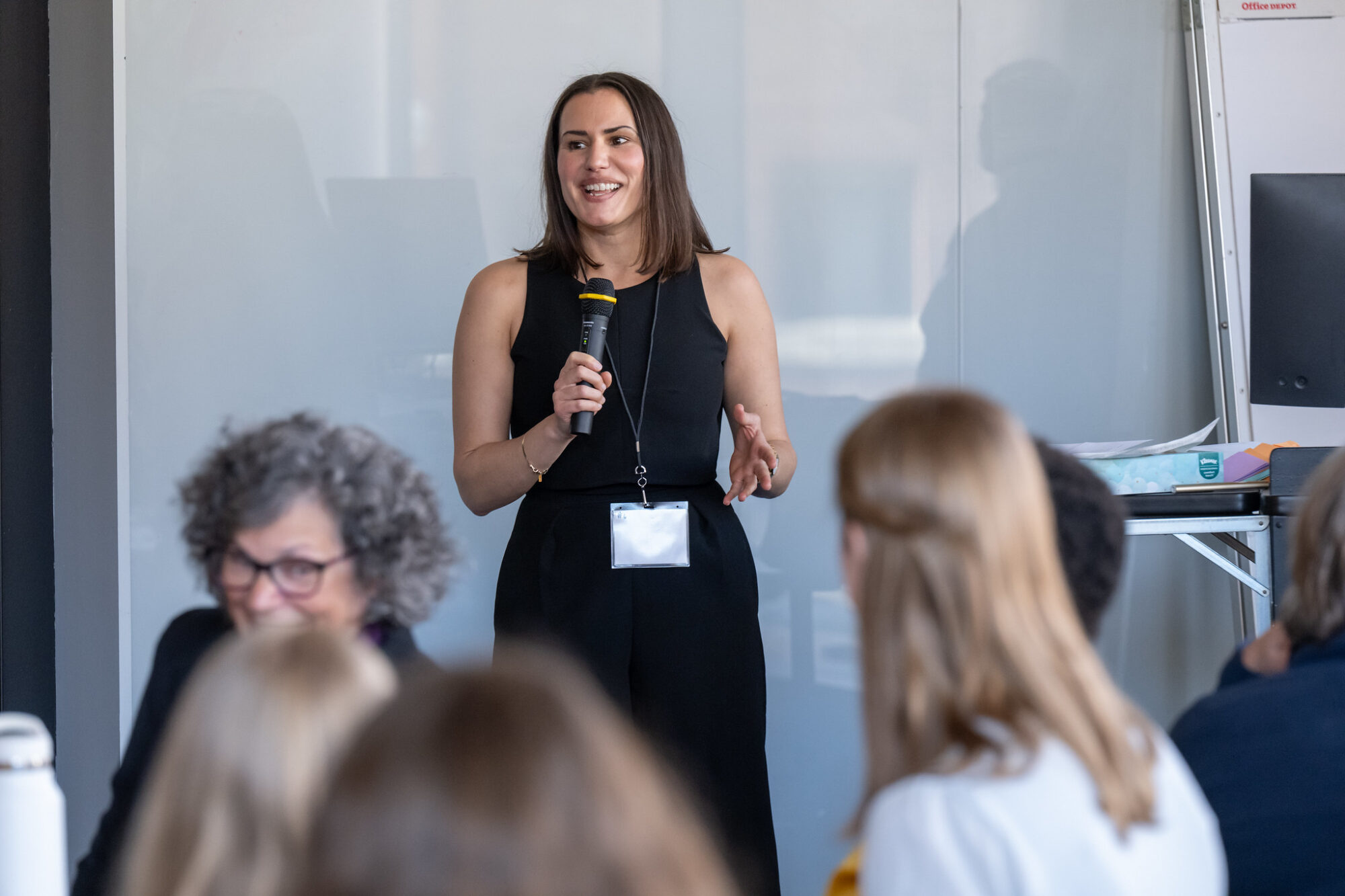 A woman stands speaking to a group, engaged in discussion, with a modern setting in the background.