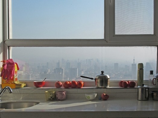 A photo showing a sunny cityscape view from a kitchen's window. On the kitchen table and near the window are some apples, a metallic pot with cover, some kitchen gloves, a red bowl and a green thermos.