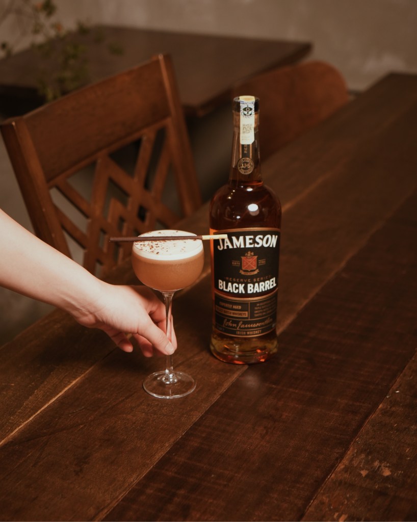 A hand holding a cocktail garnished with chocolate on a table next to a bottle of Jameson Black Barrel whiskey.