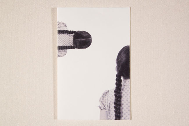 A black and white photograph shows a person from the back, with their long dark braids visible, looking towards a white background. The person’s shoulders and upper back are visible, wearing a light-colored garment with a small, repeating pattern. The image is framed by a thin white border and is presented against a light beige or cream-colored wall.