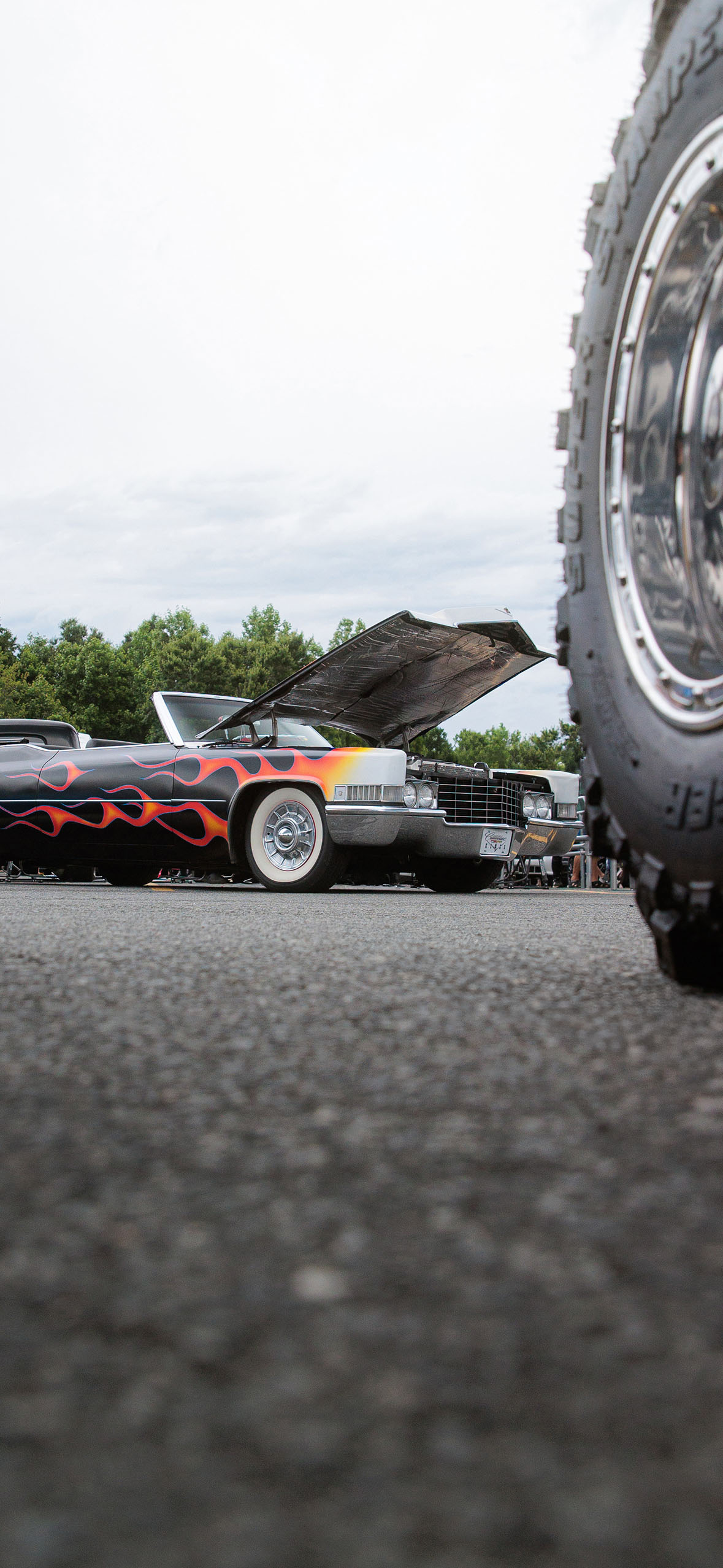 Low angle view of a vintage car with flame paint job and its hood open, partially obscured by a large tire in the foreground.