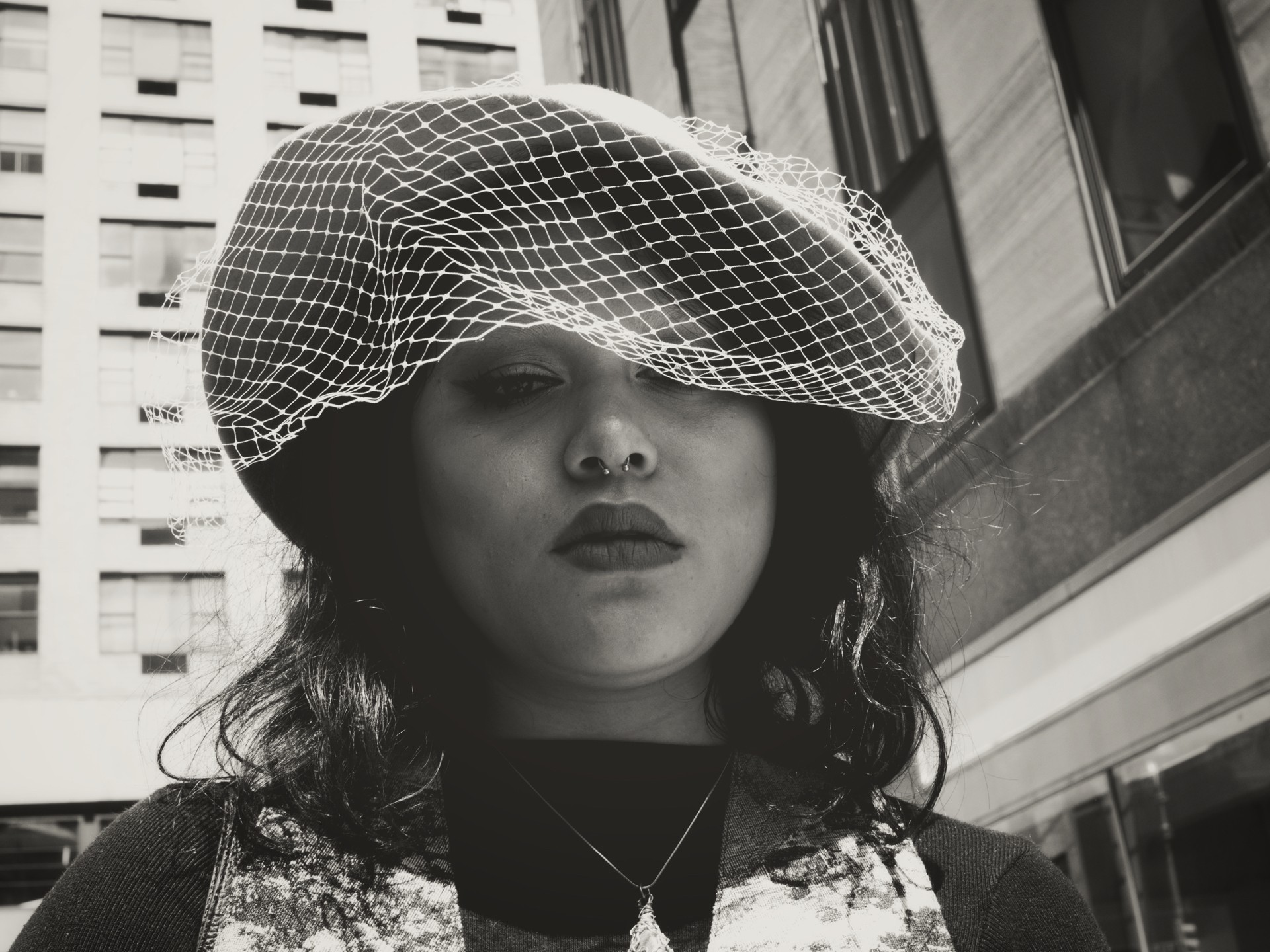 Black-and-white urban fashion portrait showing a veiled hat, bold lipstick, and high-contrast lighting against a backdrop of tall city architecture — ideal for street photography, vintage style, and editorial fashion content.