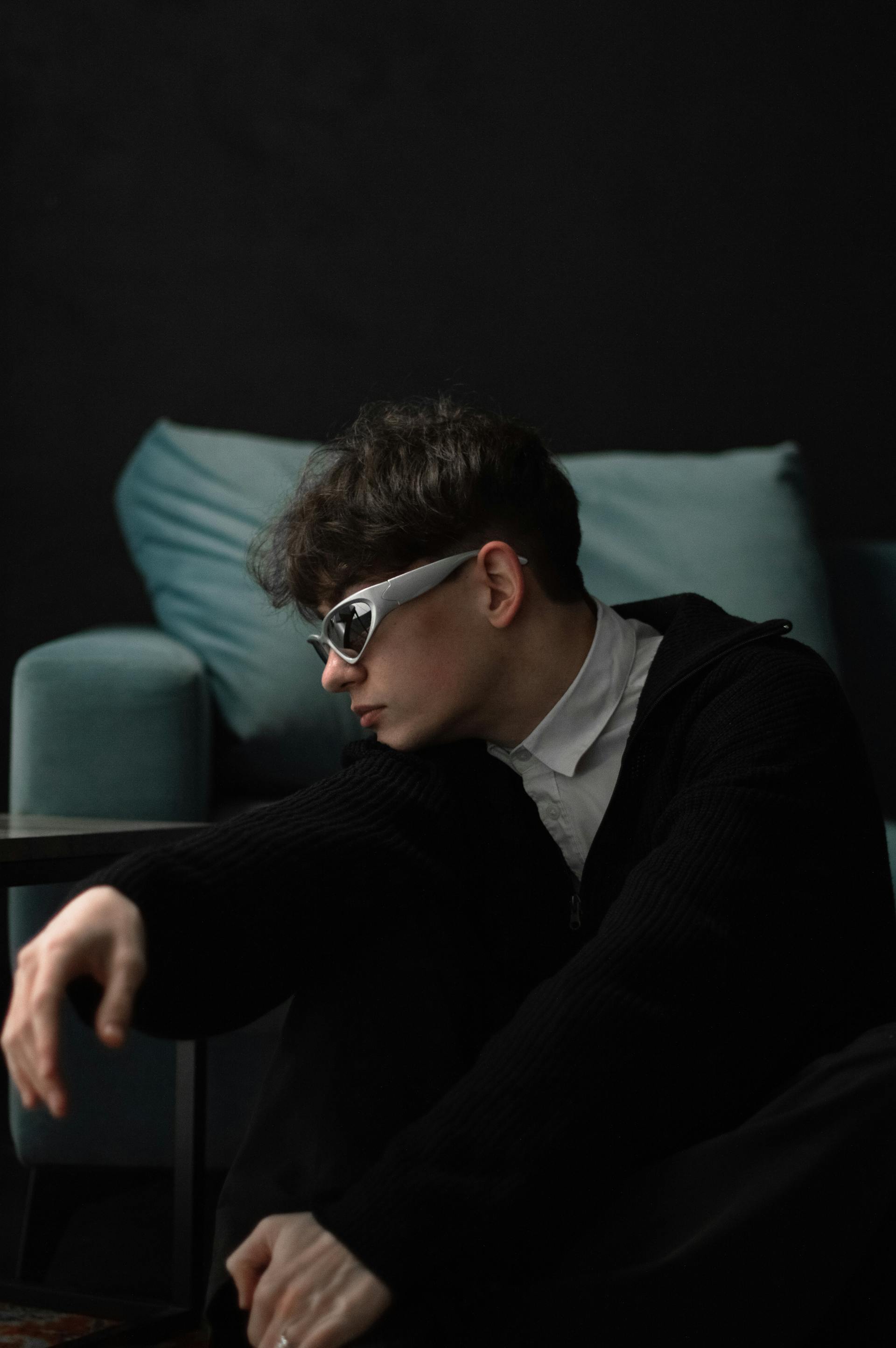 A side-profile of a young person with dark, wavy hair, wearing silver sports sunglasses and a black sweater, sitting in front of a blurred teal couch.