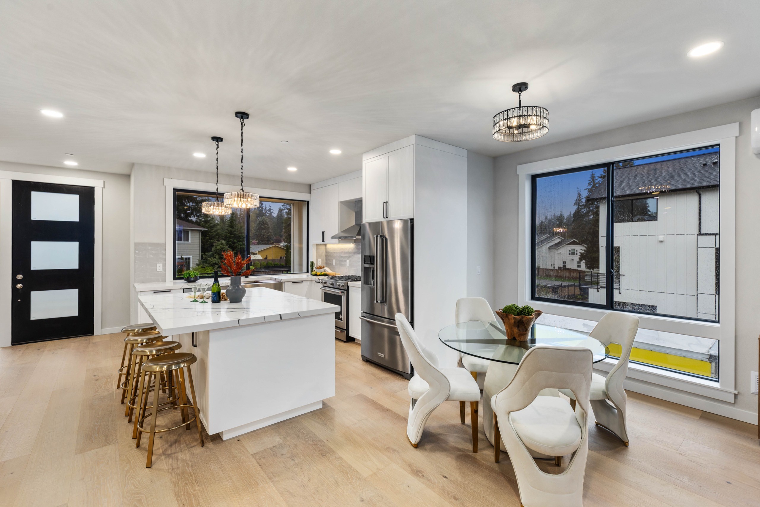 Open-plan luxury kitchen and dining area with white island, gold bar stools, glass dining table, chandelier, black-framed doors, and large picture windows
