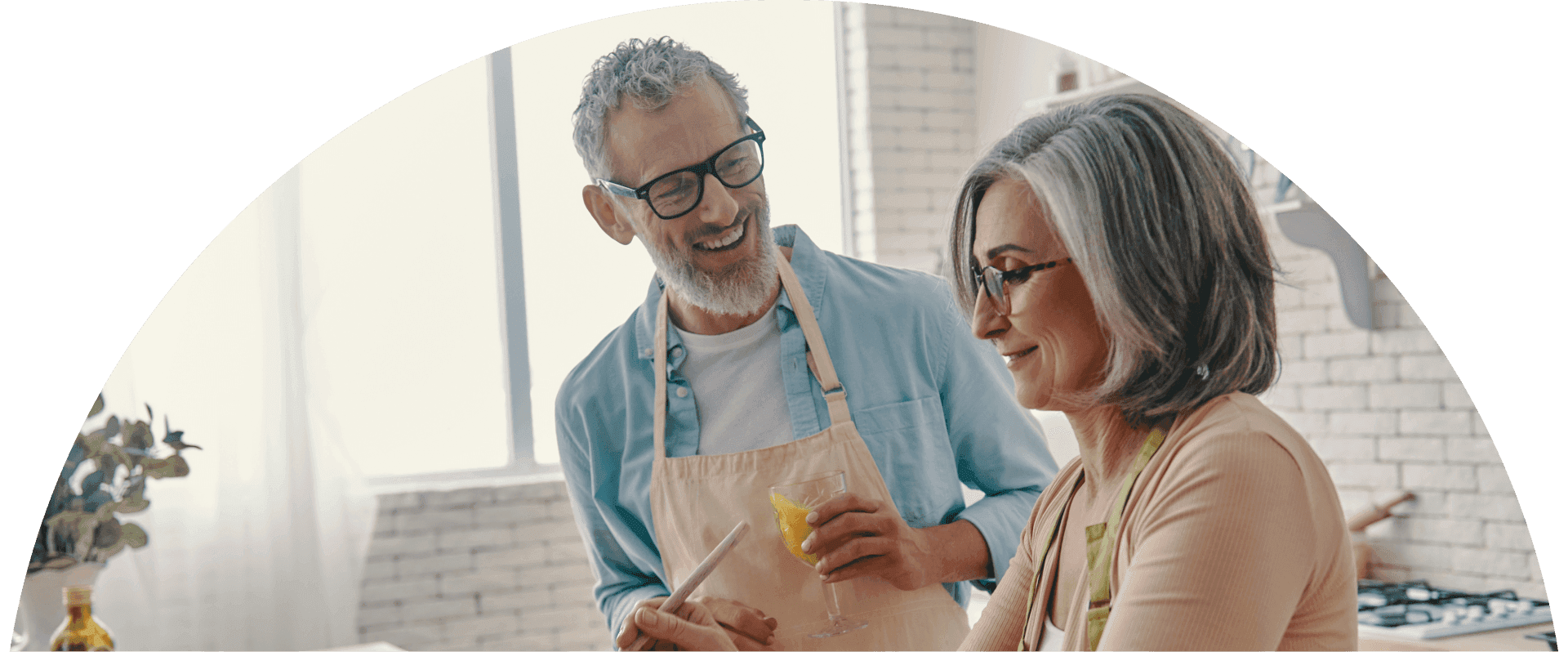 Older couple smiles while cooking together in a bright kitchen, holding a drink and preparing food