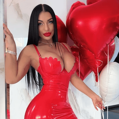 Mujer con top blanco posando frente a un auto blanco y una casa de fondo en un ambiente exterior soleado.