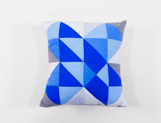 A square throw pillow with a geometric pattern rests on a white surface. The pillow features alternating blue and gray triangular shapes arranged in a symmetrical design. The background is plain white.