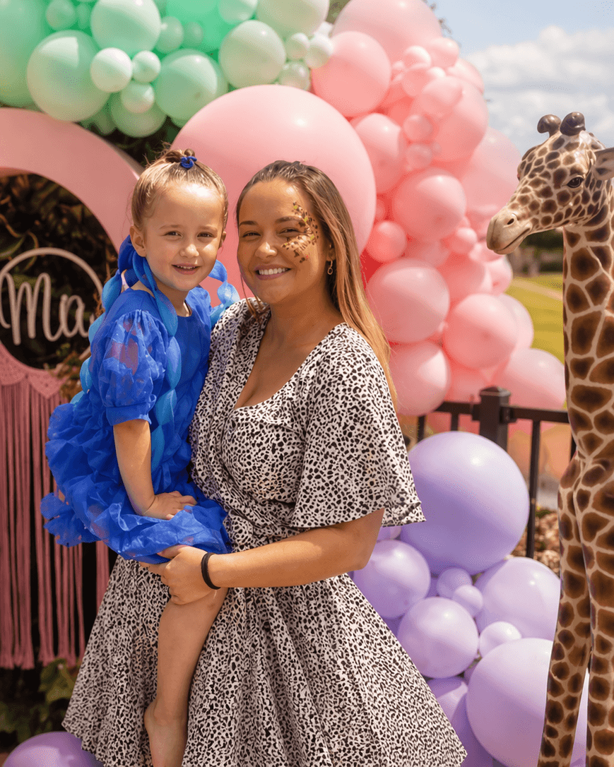 A smiling woman in a patterned dress holds a young girl in a blue dress amid pink,  green