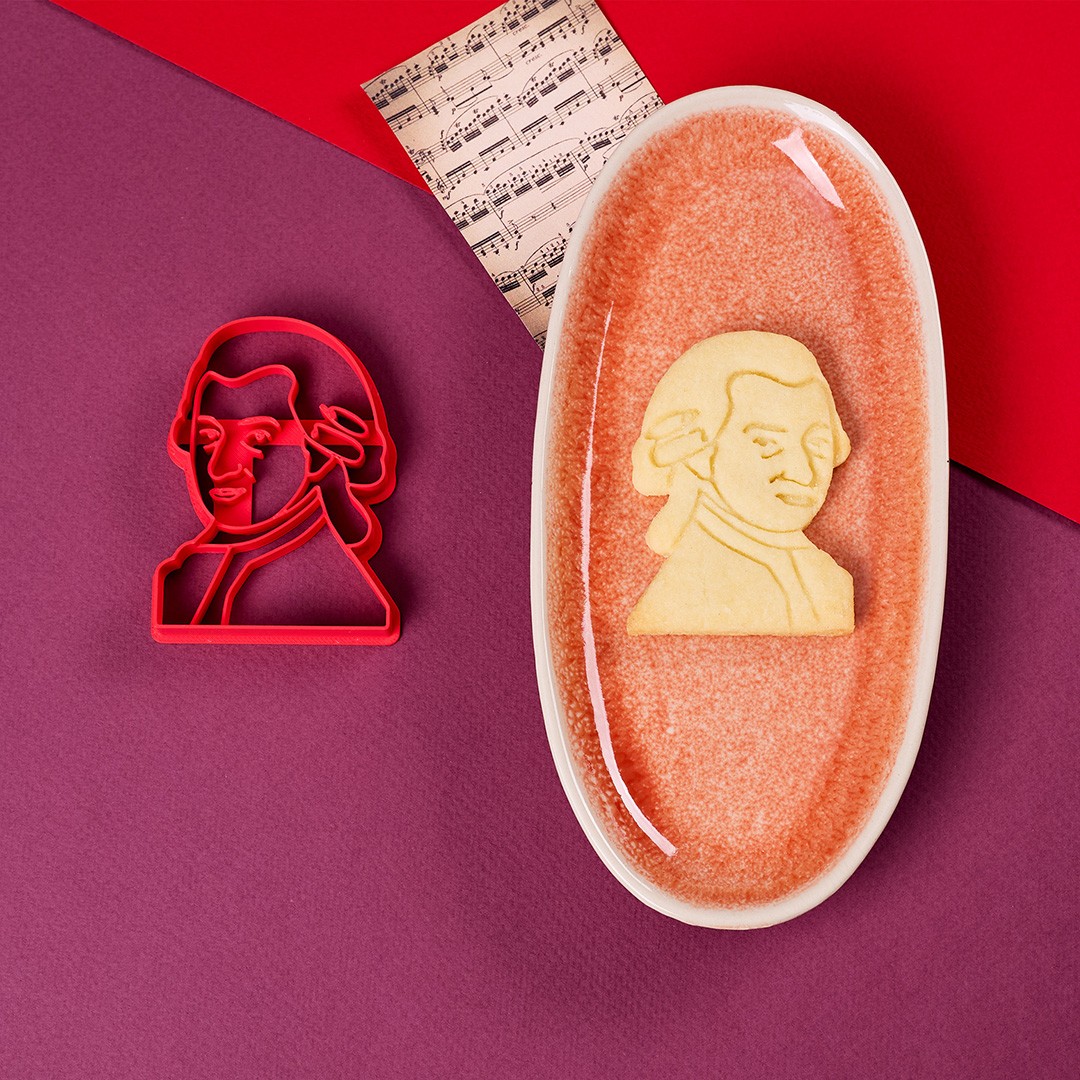 The image features a Mozart-shaped cookie cutter beside a matching cookie, set on a textured purple surface with a background of red paper and a music sheet, epitomizing Biocraftlab's creative baking tools.