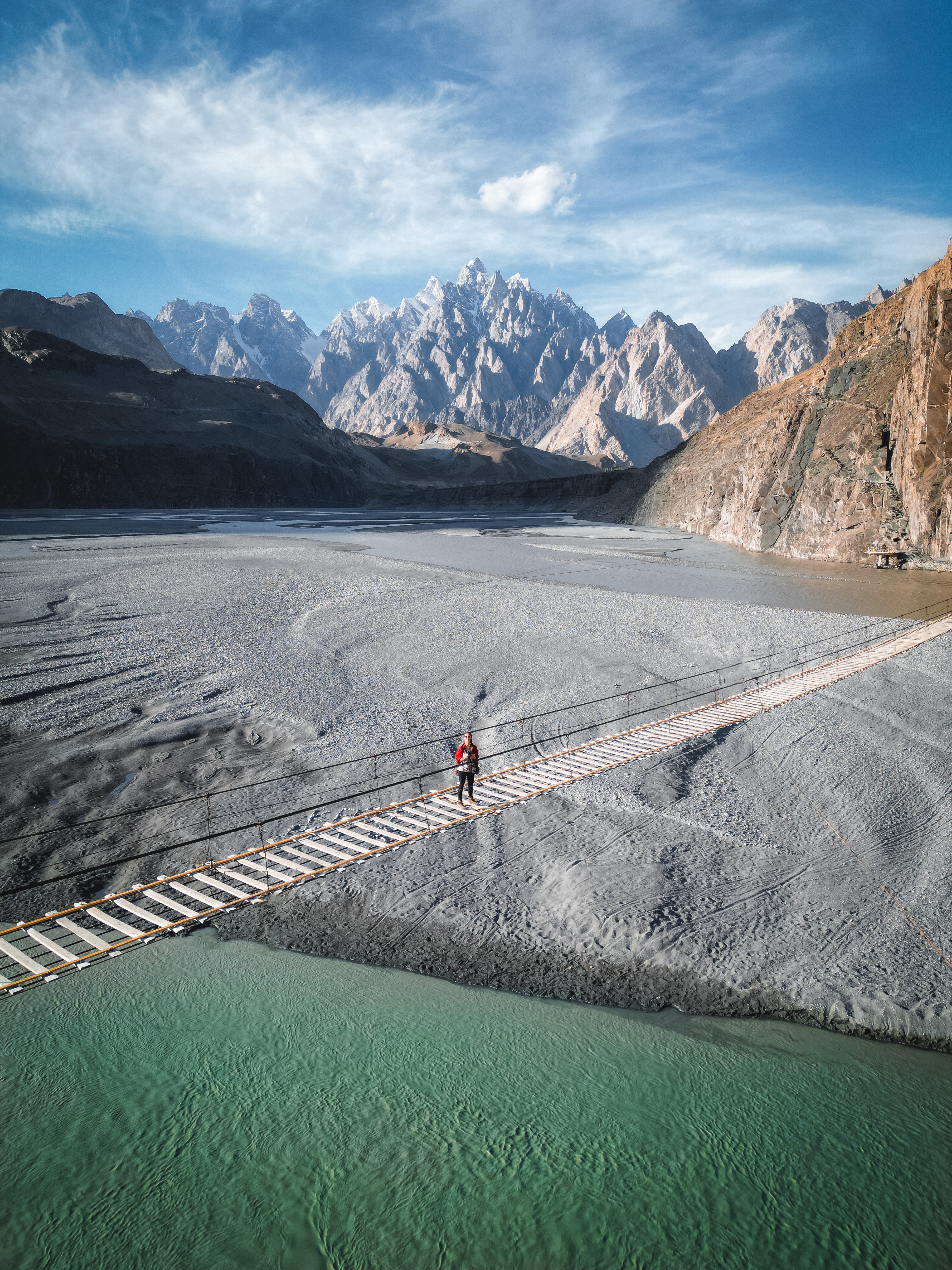 A woman stands alone in the middle of a long slatted bridge with pointy, snow-capped mountains in the background