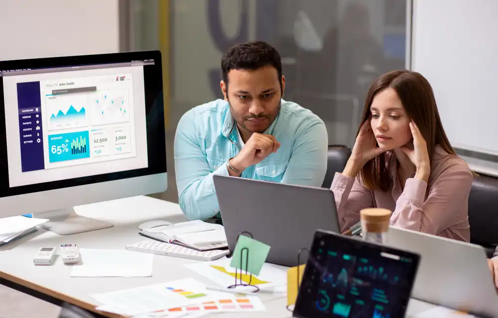A man and a woman in an office, seated beside each other at a desk with laptops and a large monitor, both look stressed, with hands to their faces.