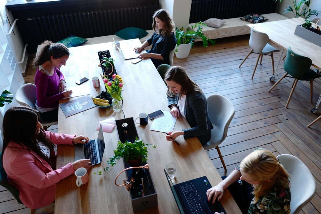 Coworking space with women collaborating around a wooden table, laptops, and plants.