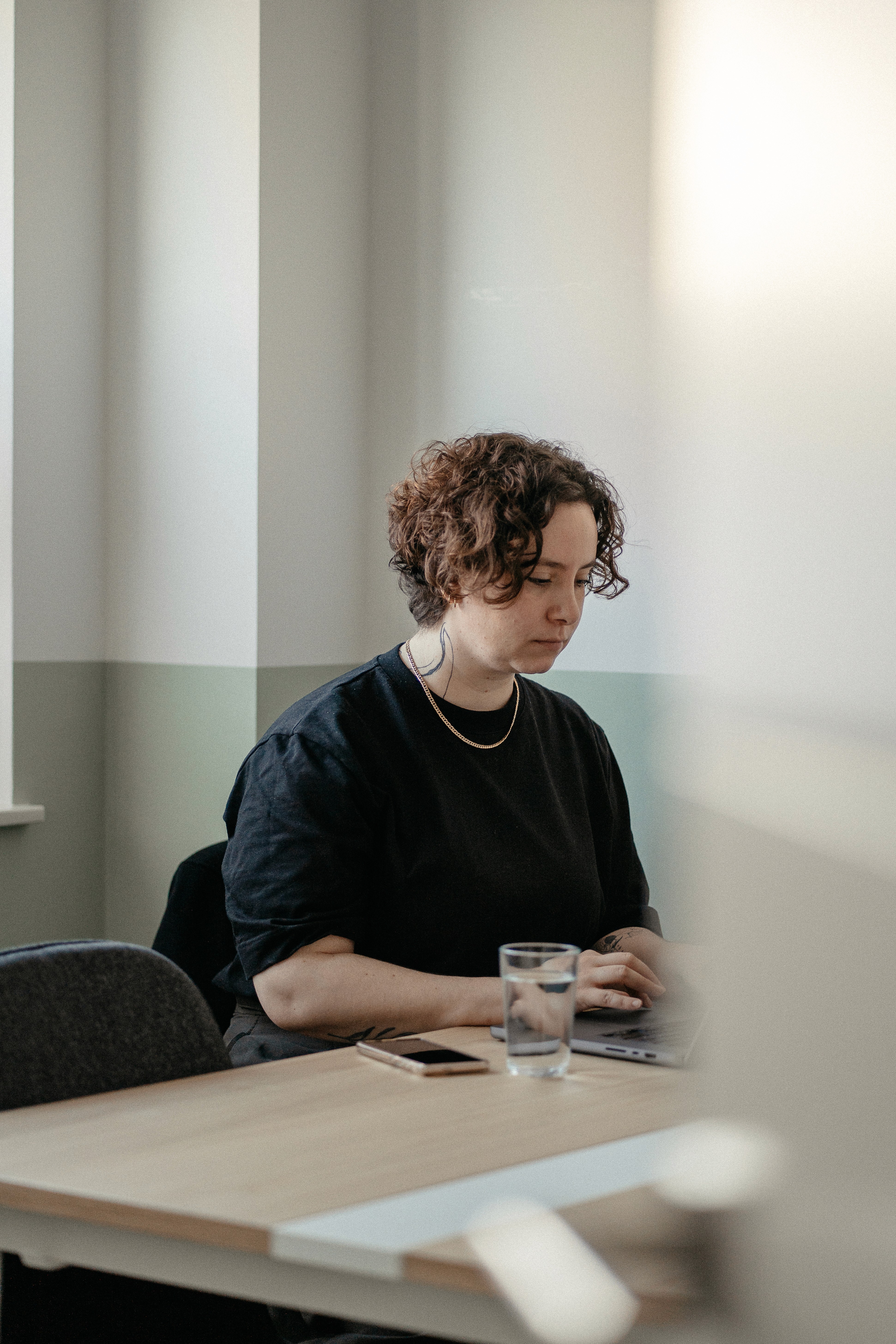 Person mit lockigem Haar und schwarzem Pullover sitzt an einem Tisch in einem Coworking-Space mit Laptop, Glas Wasser und Smartphone