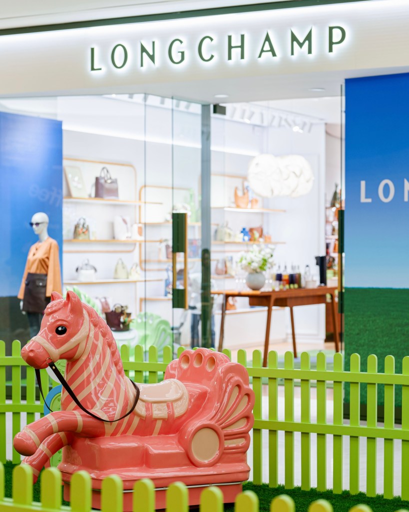 A colorful pink rocking horse in front of the Longchamp store, surrounded by a green picket fence.