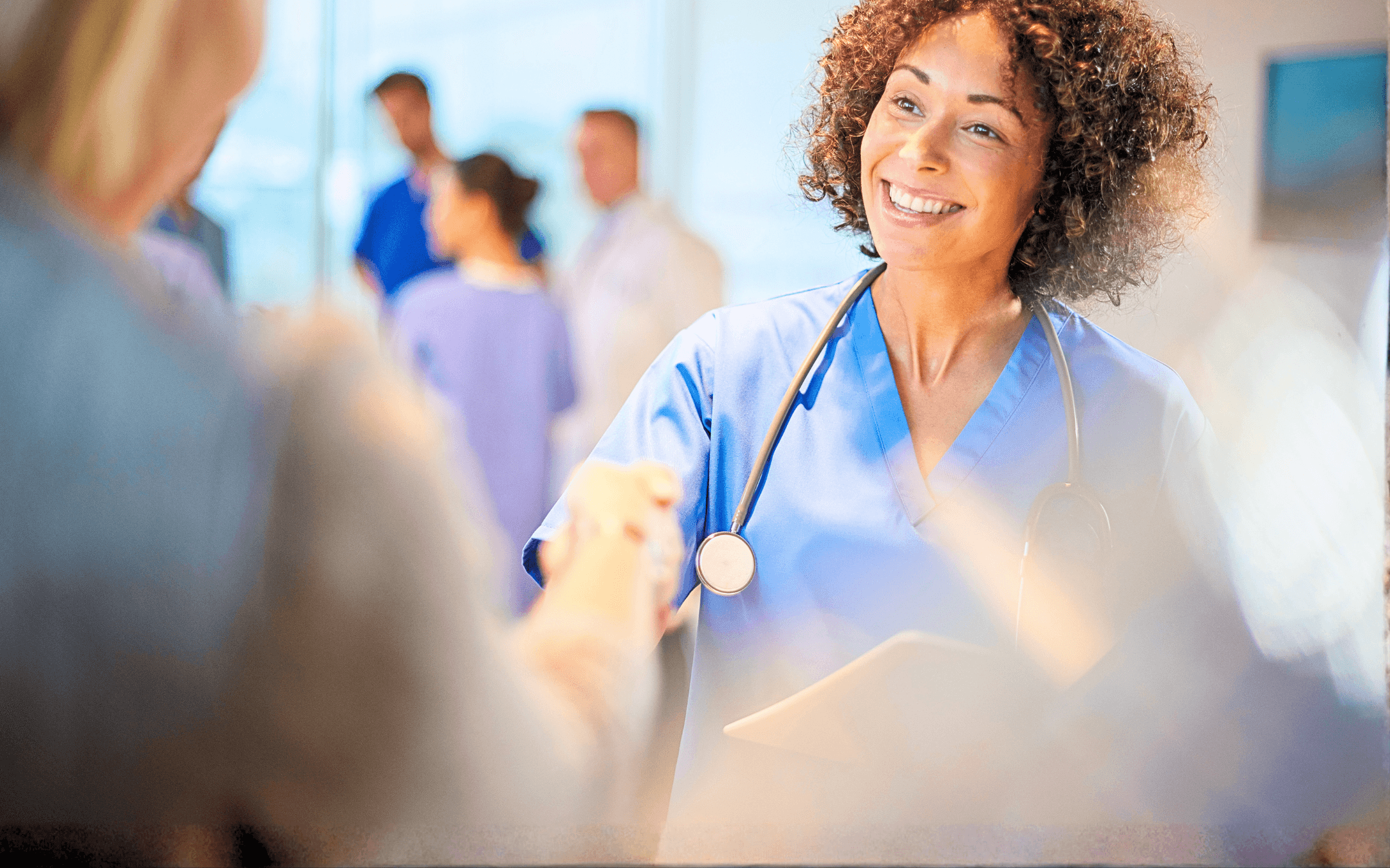 Smiling nurse shakes a patient's hand. Other medical professionals in scrubs stand in the background.