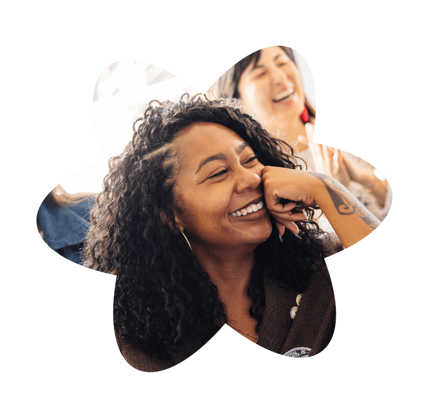 Woman with curly hair smiling and resting her chin on her hand while laughing with others in a casual group setting.