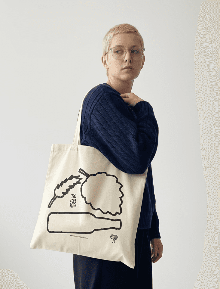 A blong young lady who has a short hair wearing a dark blue knitted jumper. On her sholder we see a custom designed tote bag with quirky illustrations of a bottle, a hop plant and a barley. 